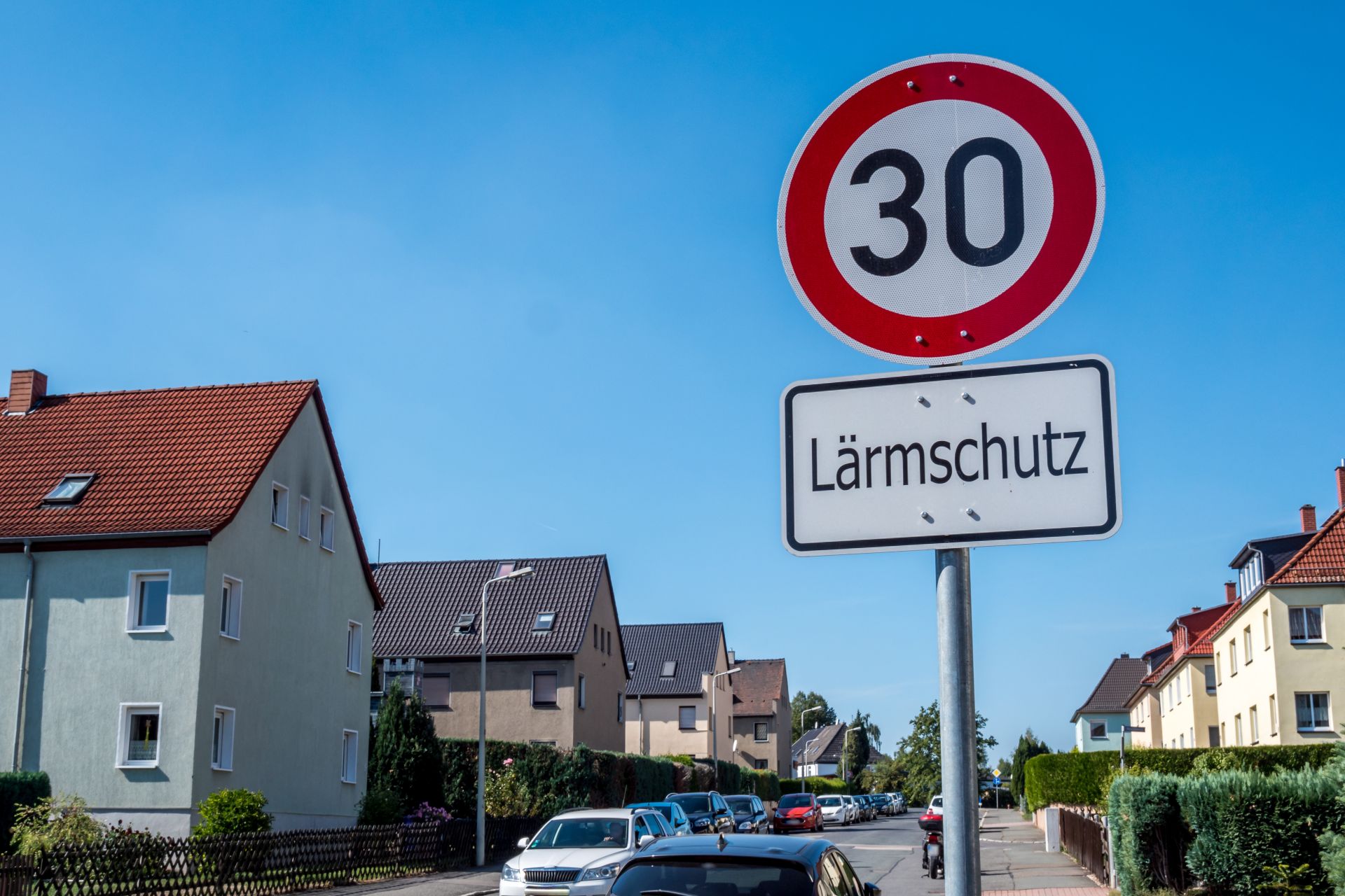 Straßenschild Geschwindigkeitsbegrenzung auf 30km/h mit Zusatzschild "Lärmschutz" in Wohngebietsstraße vor blauem Himmel.