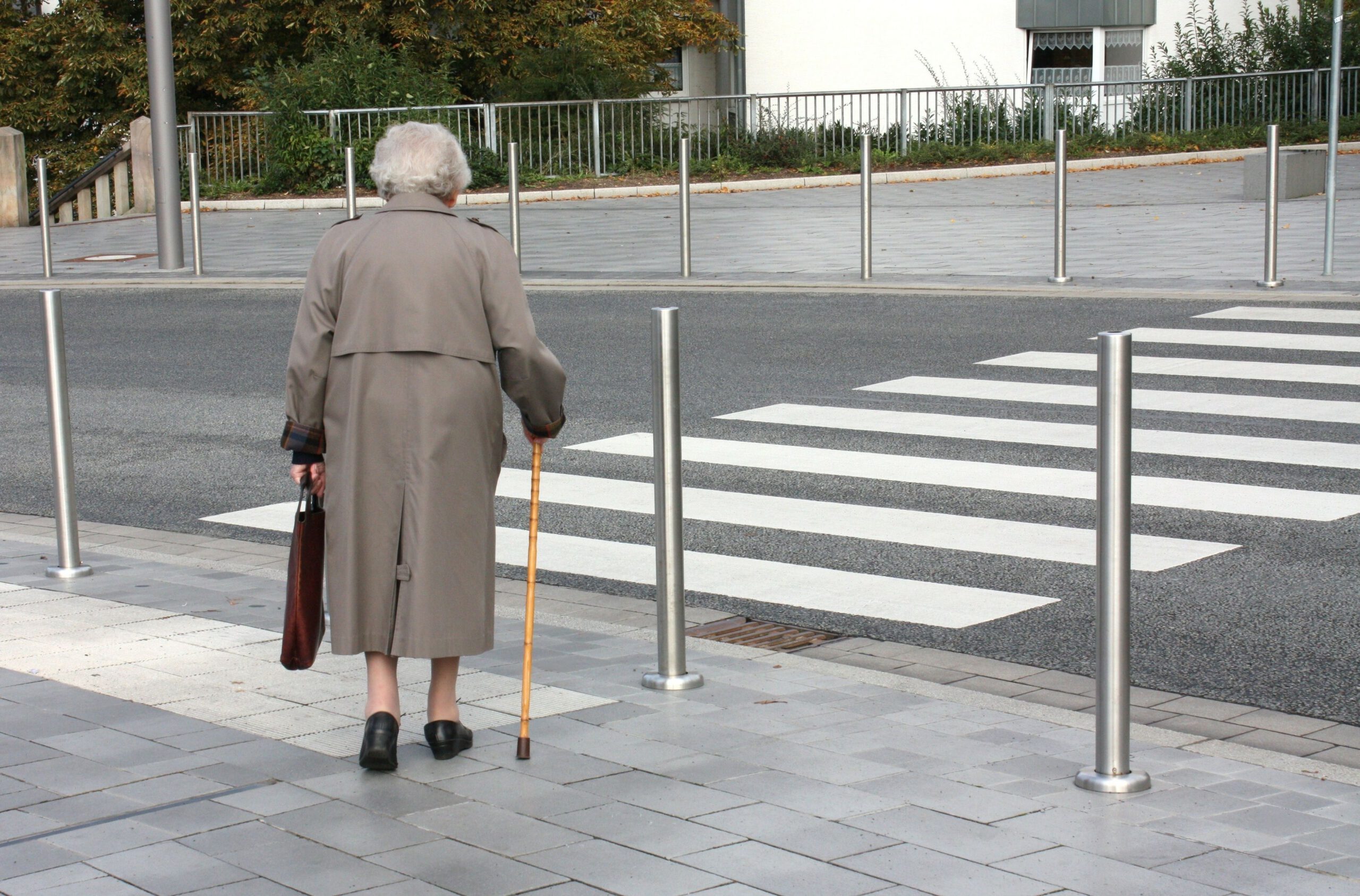 Ältere Person in grauer Jacke am Stock auf dem Gehweg zum Zebrastreifen.